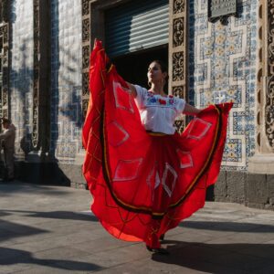 A woman dances gracefully in a vibrant red dress in front of ornate Mexican architecture.